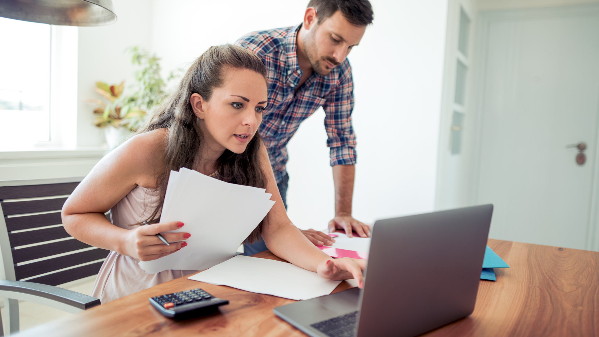 A woman sat at a table, with a man stood behind her, looking at a laptop while holding a pile of papers